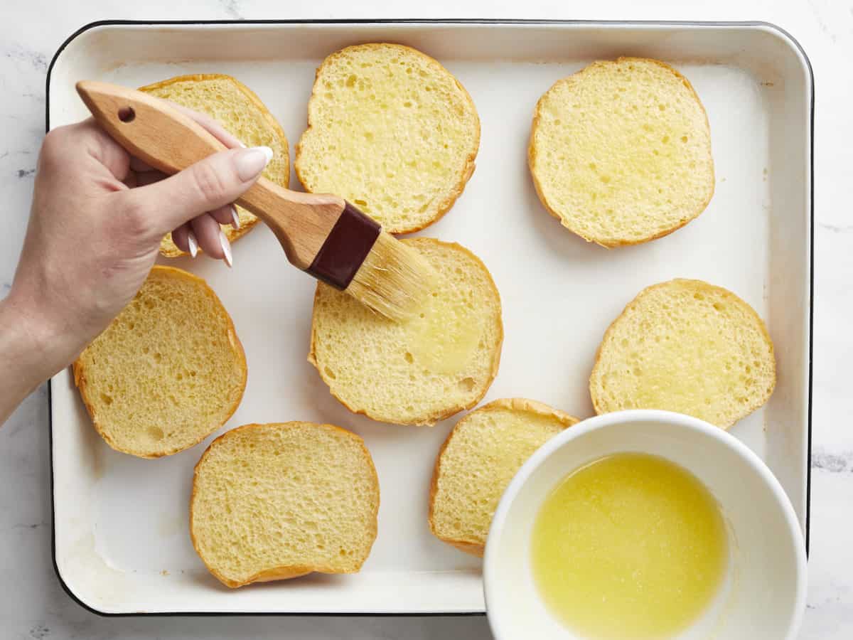 Overhead shot of melted butter being brushed onto a bun in a sheet pan with other buns.