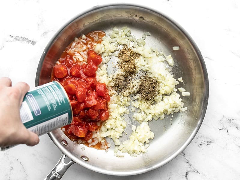 Diced tomatoes being poured into the skillet, with basil, oregano, and pepper.