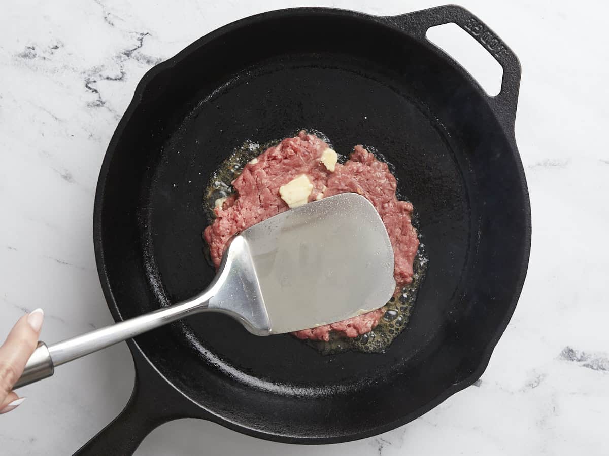 Overhead shot of raw Smashed Burger patty in a black cast iron pan being smashed by a silver spatula.