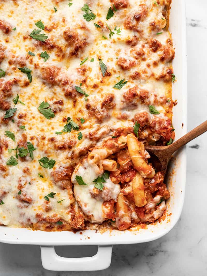 Overhead view of a wooden spoon scooping baked ziti out of the corner of a white casserole dish