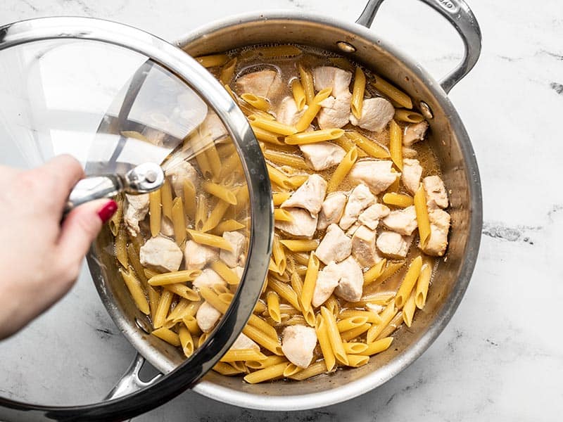 Lid being placed on the skillet with uncooked pasta and chicken