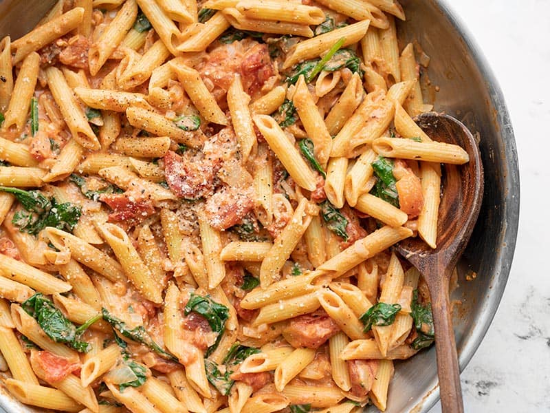 Close up overhead view of creamy tomato spinach pasta in a skillet, a wooden spoon in the side