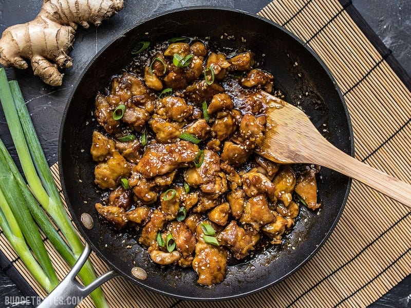 Finished easy sesame chicken in a skillet with a wooden spatula on a bamboo mat with green onion and ginger near by.