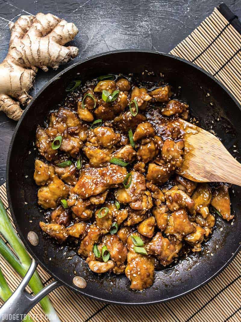 A skillet full of easy sesame chicken with a wooden spatula on a bamboo mat.