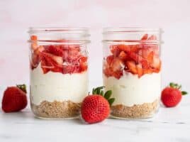 Two jars of no bake strawberry cheesecake side by side with whole strawberries on the side