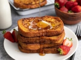 Side view of four slices of french toast stacked on a white plate being drizzled with maple syrup.