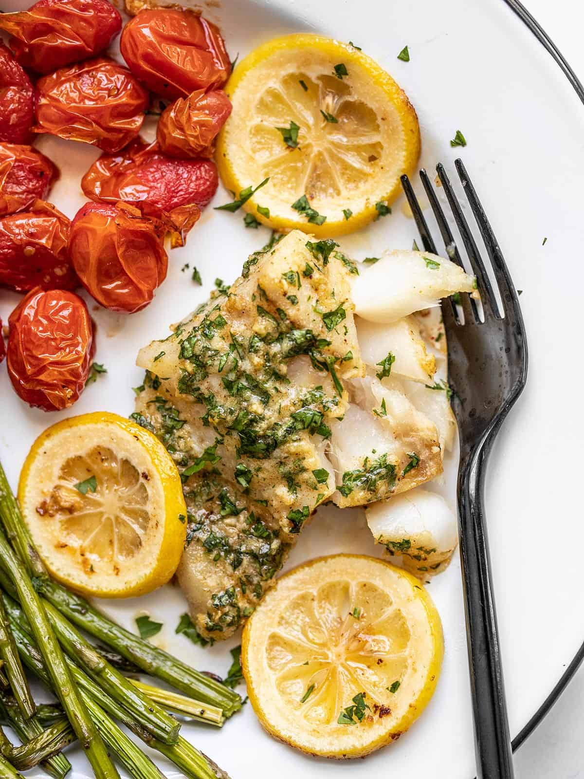 garlic butter baked cod on a plate with tomatoes, asparagus, and a fork