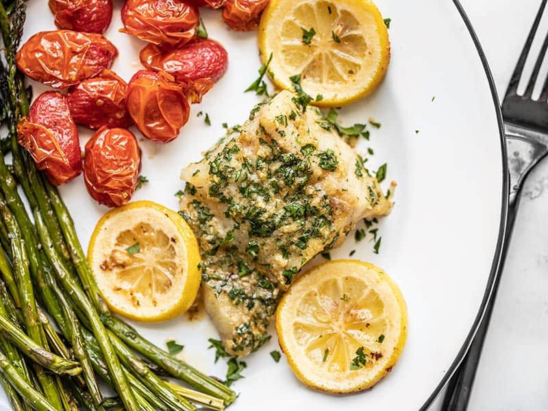 Overhead view of Garlic Butter Baked Cod on a plate with tomatoes and asparagus, a black fork on the side.