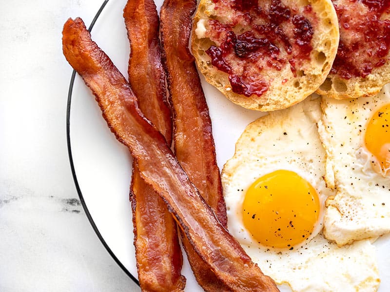 Three strips of bacon on a plate with fried eggs and an English Muffin