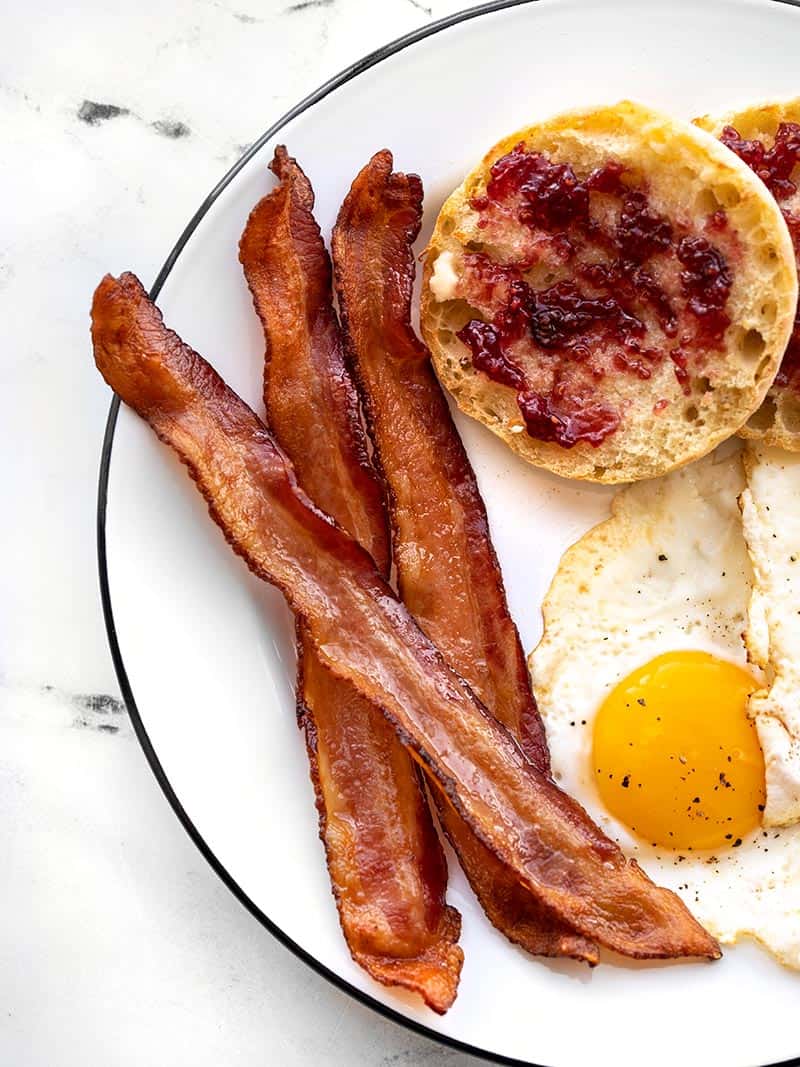Three strips of cooked bacon on a plate with fried eggs and an English muffin with jam
