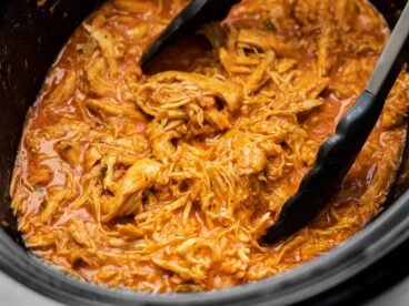 Close up side view of shredded buffalo chicken in the slow cooker with tongs