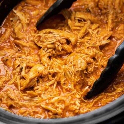 Close up side view of shredded buffalo chicken in the slow cooker with tongs