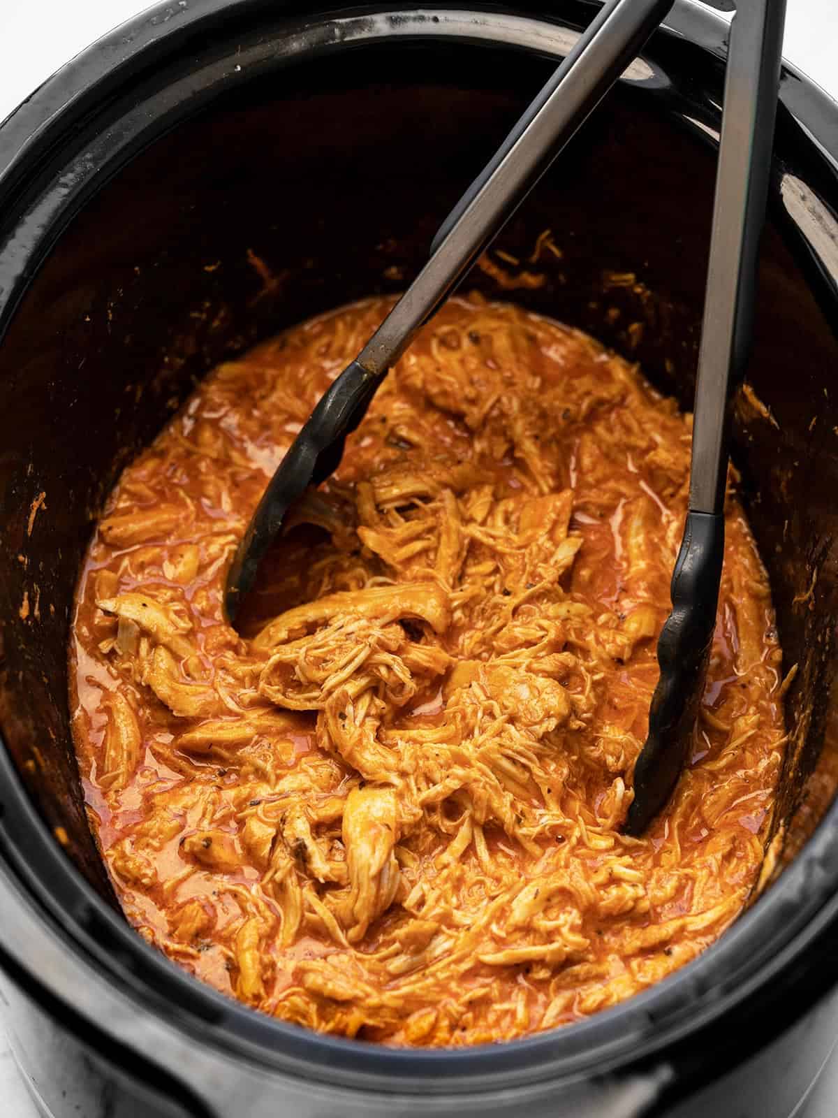 Shredded buffalo chicken in a slow cooker with tongs