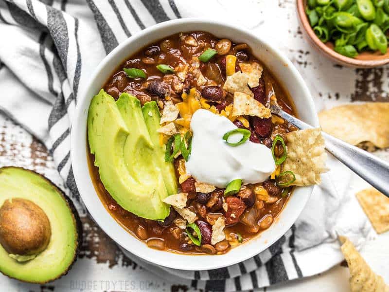 A close up view of a bowl of Slow Cooker Vegetarian Lentil Chili fully dressed with avocado, sour cream, cheese, and chips