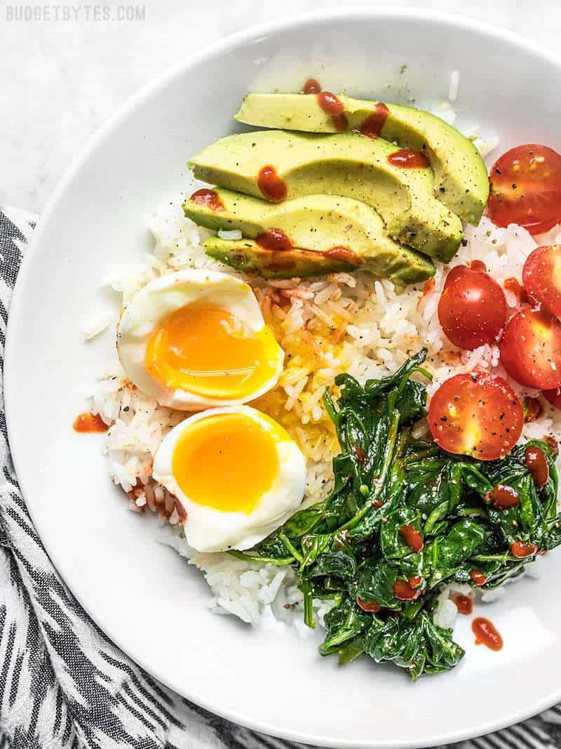 Breakfast bowl with perfectly cooked soft boiled eggs, spinach, avocado, tomato, rice, and sriracha.