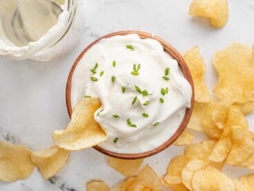 Overhead shot of a wooden bowl with sour cream in it that's topped with chives and is surrounded by potato chips.